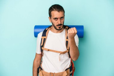 Mavi arka planda izole edilmiş genç beyaz adam yumruğunu kameraya gösteriyor, agresif yüz ifadesi..