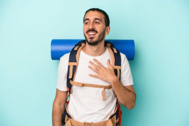 Mavi arka planda izole edilmiş genç beyaz yürüyüşçü elini göğsünde tutarak kahkahalar atıyor..