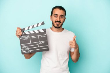 Kafkasyalı genç aktör elinde alkış tahtası tutarken mavi arka planda izole edilmiş gülümsüyor ve başparmağını kaldırıyor.