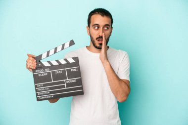 Beyaz tenli genç bir aktör mavi arka planda izole edilmiş bir şekilde el çırpma tahtası tutuyor.