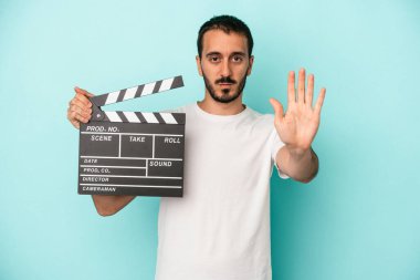 Beyaz tenli genç aktör elinde alçıpanla mavi arka planda izole edilmiş, elini uzatmış, dur işaretini göstererek seni engelliyor..