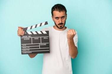 Kafkasyalı genç aktör elinde klaket tutarken mavi arka planda kameraya yumruk ve agresif yüz ifadesini gösteriyor..