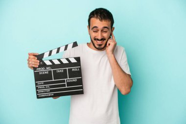 Kafkasyalı genç aktör elinde alkış tahtası tutarken mavi arka planda kulakları elleriyle kapatıyor..