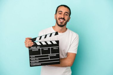 Kafkasyalı genç aktör elinde klaket tutarken mavi arka planda izole edilmiş gülüyor ve eğleniyor..