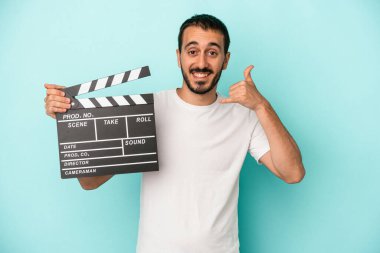 Kafkasyalı genç aktör elinde klaket tutarken mavi arka planda parmaklarıyla bir cep telefonu hareketi gösteriyor..