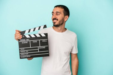 Kafkasyalı genç aktör elinde alkış tutarken mavi arka planda tek başına gülümsüyor, neşeli ve keyifli görünüyor..