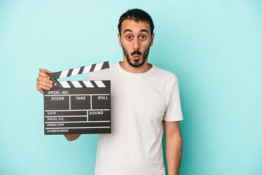 Kafkasyalı genç aktör elinde alkış tahtası, mavi arka planda izole edilmiş omuzlar ve açık gözler..