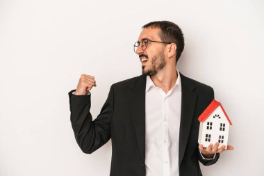 Genç emlakçı, beyaz arka planda izole edilmiş bir model evi tutuyor. Zaferden sonra yumruğunu kaldırıyor..