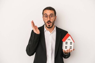 Genç emlakçı elinde model bir ev tutuyor. Beyaz arka planda izole edilmiş. Şaşırmış ve şok olmuş..