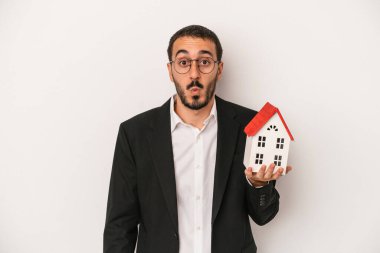 Genç emlakçı elinde beyaz arka planda izole edilmiş bir ev tutuyor omuzlarını silkiyor ve gözleri açık..