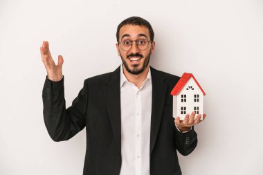 Genç emlakçı, beyaz arka planda izole edilmiş bir model evi tutuyor. Hoş bir sürprizle karşılaşıyor, heyecanlı ve el kaldırıyor..