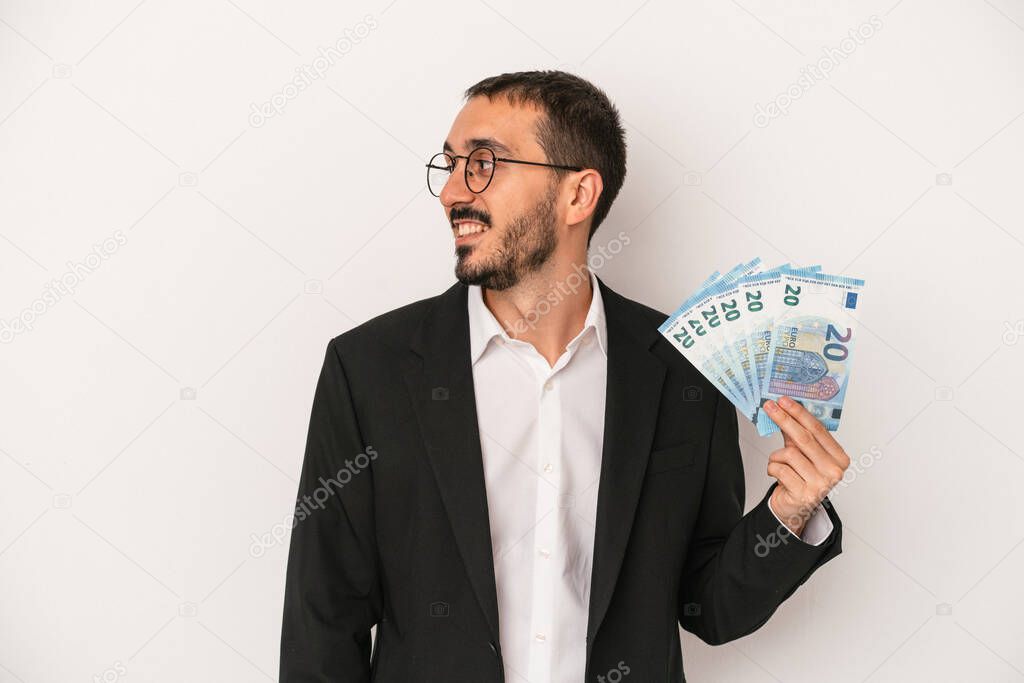 Young caucasian business man holding banknotes isolated on white background looks aside smiling, cheerful and pleasant.