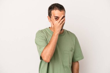 Beyaz arka planda izole edilmiş genç beyaz bir adam kameraya parmaklarıyla göz kırpıyor, yüzünü kapatmaktan utanıyor..