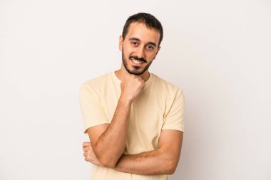 Beyaz arka planda izole olmuş genç beyaz bir adam gülümsüyor ve kendinden emin, çenesine elleriyle dokunuyor..