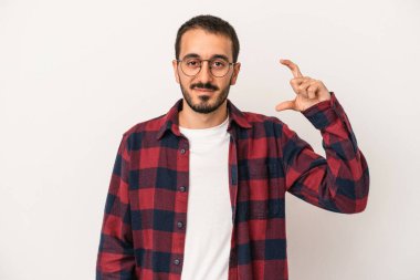 Beyaz arka planda izole edilmiş genç beyaz bir adam işaret parmaklarıyla küçük bir şey tutuyor, gülümsüyor ve kendinden emin..