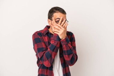 Beyaz arka planda izole edilmiş genç beyaz bir adam parmaklarının arasından korkmuş ve gergin bir şekilde göz kırpıyor..