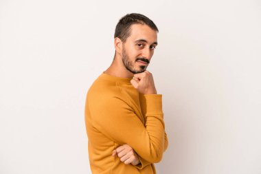 Beyaz arka planda izole olmuş genç beyaz bir adam gülümsüyor ve kendinden emin, çenesine elleriyle dokunuyor..