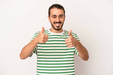 Beyaz arka planda izole edilmiş genç beyaz adam gülümsüyor ve başparmağını kaldırıyor.