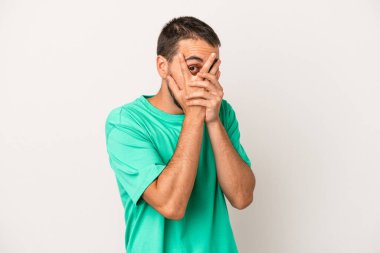 Beyaz arka planda izole edilmiş genç beyaz bir adam parmaklarının arasından korkmuş ve gergin bir şekilde göz kırpıyor..