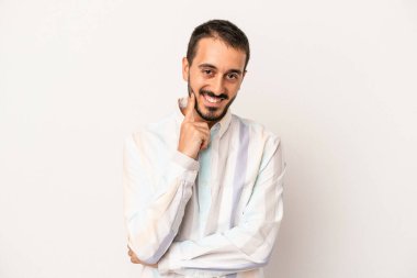 Beyaz arka planda izole olmuş genç beyaz bir adam gülümsüyor ve kendinden emin, çenesine elleriyle dokunuyor..