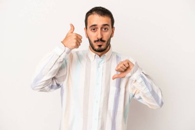 Beyaz arka planda izole edilmiş genç beyaz bir adam baş parmaklarını aşağı ve yukarı kaldırıyor, zor seçim konsepti