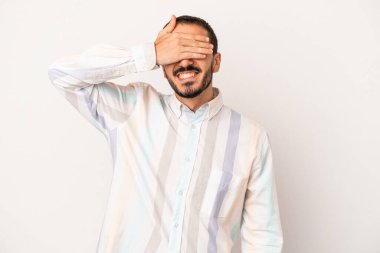 Beyaz arka planda soyutlanmış beyaz tenli genç bir adam elleriyle gözlerini kapatıyor, geniş bir gülümsemeyle bir sürpriz bekliyor..