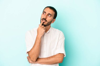 Mavi arka planda izole edilmiş genç beyaz adam şüpheli ve şüpheci bir ifadeyle yana bakıyor..