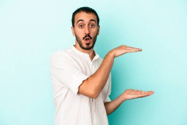Mavi arka planda izole genç beyaz adam şok ve hayret içinde elleri arasında bir kopya boşluk tutuyor.