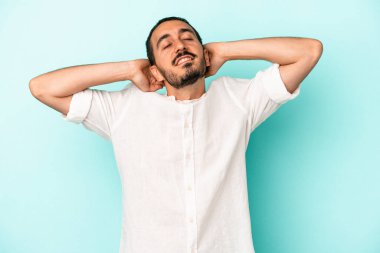 Kafkasyalı genç adam mavi arka planda izole edilmiş. Kendine güveniyor, elleri başının arkasında..