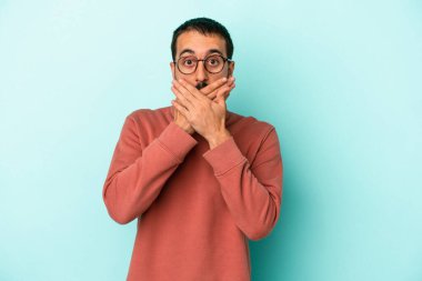 Mavi arka planda izole edilmiş genç beyaz adam elleriyle ağzını kapatırken şok oldu..