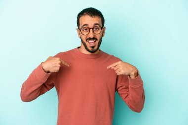 Mavi arka planda izole edilmiş genç beyaz adam parmağını göstererek şaşırdı, geniş bir gülümsemeyle..