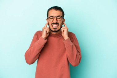 Mavi arka planda izole edilmiş genç beyaz adam kulaklarını elleriyle kapatıyor..