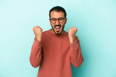 Kafkasyalı genç adam mavi arka planda izole edilmiş kaygısız ve heyecanlı tezahüratlar yapıyor. Zafer konsepti.