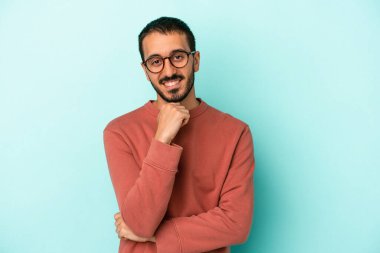 Mavi arka planda izole edilmiş genç beyaz adam gülümsüyor ve kendinden emin, elleriyle çenesine dokunuyor..