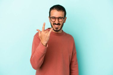 Mavi arka planda izole edilmiş genç beyaz adam parmaklarıyla kaya hareketi gösteriyor.