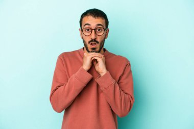 Kafkasyalı genç adam mavi arka planda izole edilmiş şans için dua ediyor, şaşırmış ve ağzını açıp önüne bakıyor..