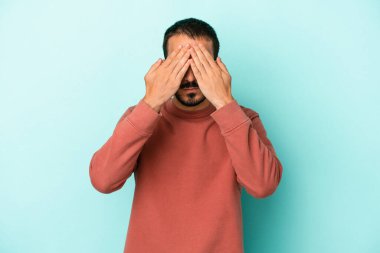 Mavi arka planda izole edilmiş genç beyaz adam elleriyle gözlerini kapatmaktan korkuyor..