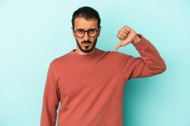 Mavi arka planda izole genç beyaz adam baş parmağını aşağı gösteriyor, hayal kırıklığı konsepti.