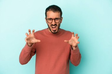 Mavi arka planda izole edilmiş genç beyaz adam pençelerini kedi taklidi yaparken gösteriyor, agresif bir hareket..