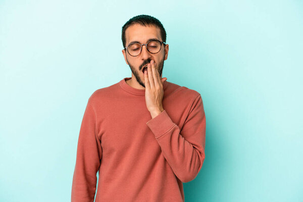 Молодой кавказский мужчина на голубом фоне зевает, показывая усталый жест, прикрывающий рот рукой..