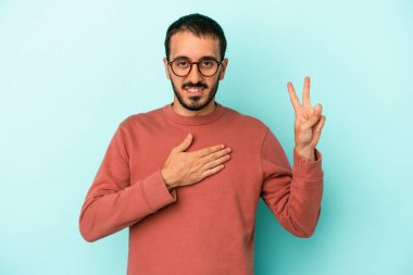 Kafkasyalı genç adam mavi arka planda izole edilmiş yemin ediyor, elini göğsüne koyuyor..