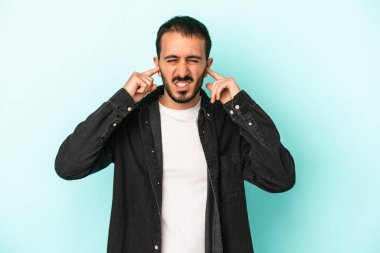 Genç beyaz adam mavi arka planda izole edilmiş parmaklarıyla kulaklarını kapatıyor, gürültülü bir ortam tarafından stresli ve umutsuz..