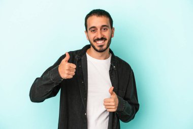 Kafkasyalı genç adam mavi arka planda izole edilmiş. Baş parmağını kaldırıyor, gülümsüyor ve kendinden emin..