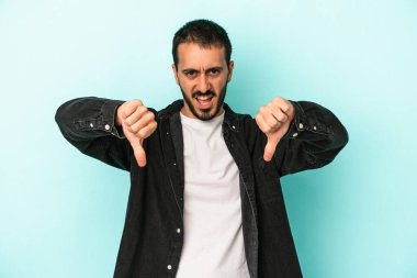 Mavi arka planda izole edilmiş genç beyaz bir adam baş parmağını aşağı gösteriyor ve hoşlanmadığını ifade ediyor..