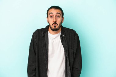 Mavi arka planda izole edilmiş genç beyaz adam omuzlarını silkeliyor ve gözleri açık..