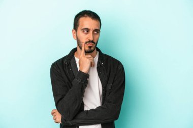 Mavi arka planda izole edilmiş genç beyaz adam şüpheli ve şüpheci bir ifadeyle yana bakıyor..