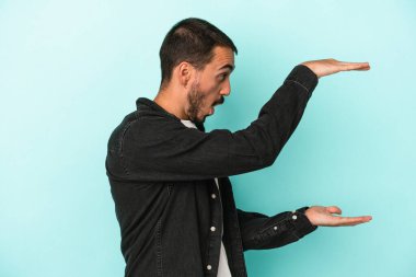 Mavi arka planda izole genç beyaz adam şok ve hayret içinde elleri arasında bir kopya boşluk tutuyor.