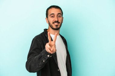 Mavi arka planda izole edilmiş genç beyaz adam zafer işareti gösteriyor ve gülümsüyor..