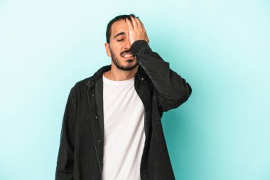 Kafkasyalı genç adam mavi arka planda izole edilmiş bir şekilde bir şeyi unutuyor. Alnına avuç içi ve gözlerini kapayarak vuruyor..