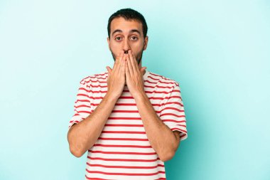 Mavi arka planda izole edilmiş genç beyaz adam elleriyle ağzını kapatırken şok oldu..